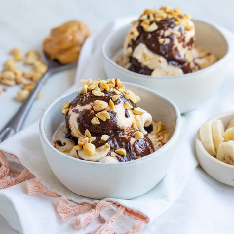 Two bowls of peanut butter ice cream and chocolate sauce topped with nuts and bananas on a white surface.