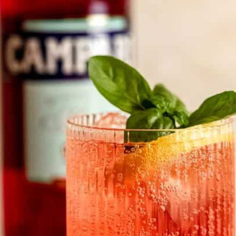 Cocktail with basil leaves and lemon slice in a glass, Campari bottle in the background