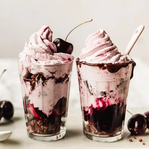 Two glasses of chocolate and cherry ice cream with cherries and fudge topping on a light background